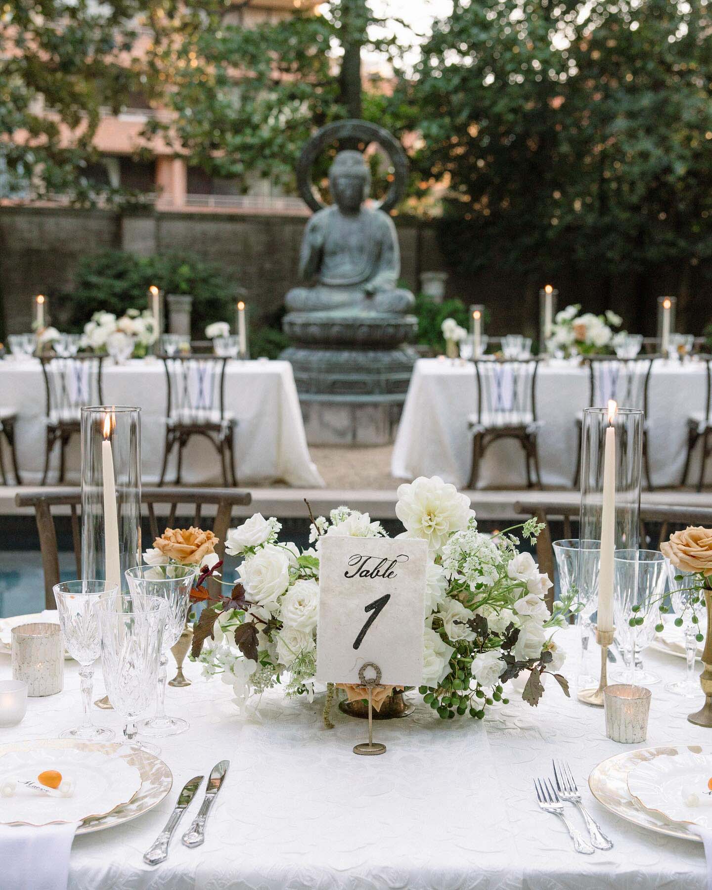 Outdoor wedding reception table featuring a white floral centerpiece, Table 1 sign, crystal glassware, taper candles, and a Buddha statue in the reflecting pool background.