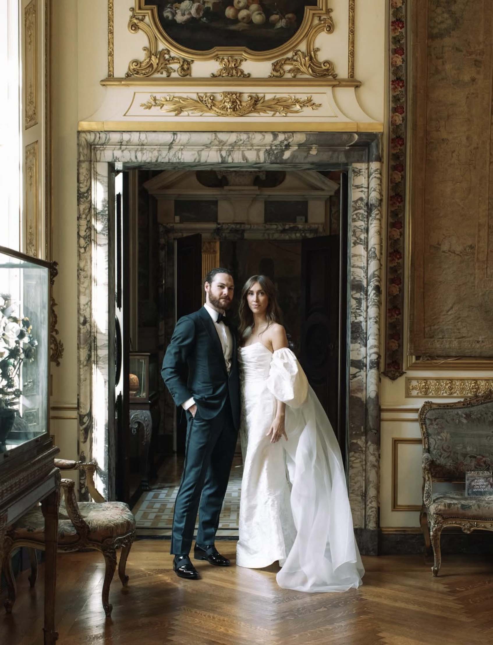 Bride and groom portrait showcasing the bride in a custom off-shoulder gown and groom in a dark green tuxedo, standing in the ornate Beaux Arts interior