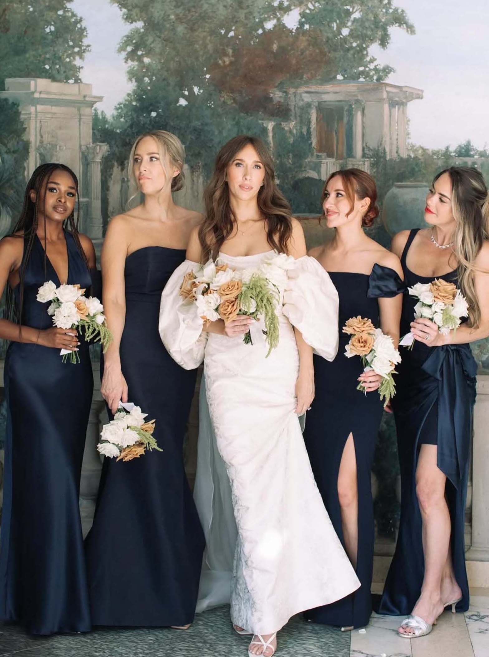 Bride in off-shoulder white gown with four bridesmaids in navy dresses posing against a Beaux Arts mural 