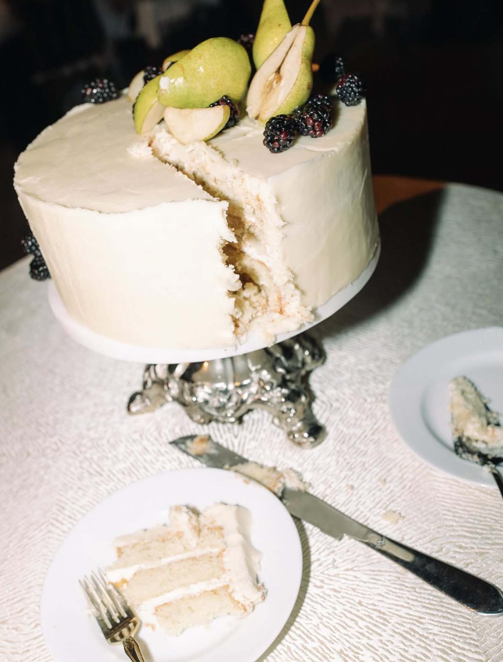 Wedding cake topped with fresh pears and blackberries, displayed on a silver stand, with a slice served on a plate.