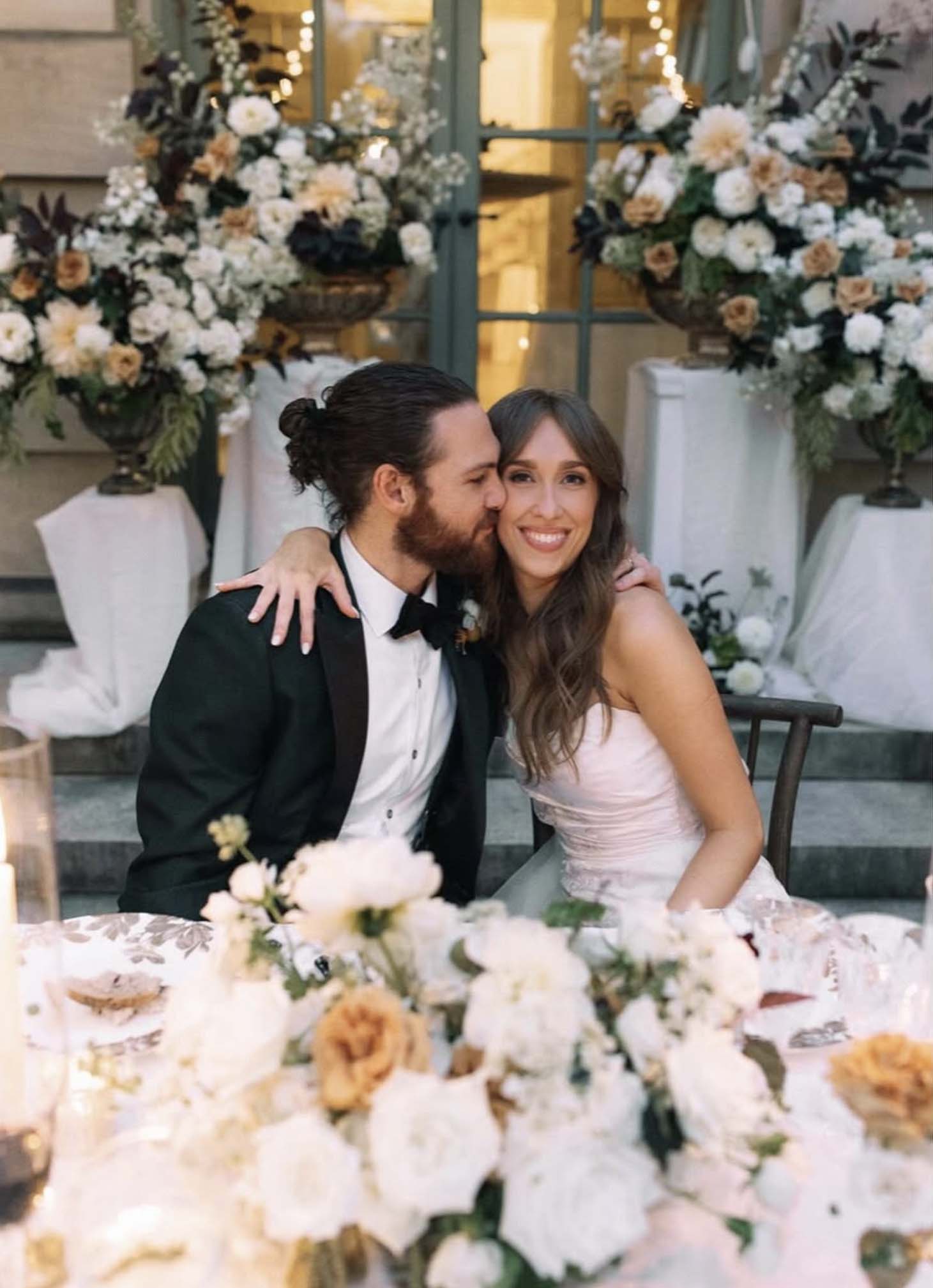 Groom in dark green tuxedo kissing bride in off-shoulder white gown as they sit at an outdoor dinner table, surrounded by lush fall floral arrangements and string lights.