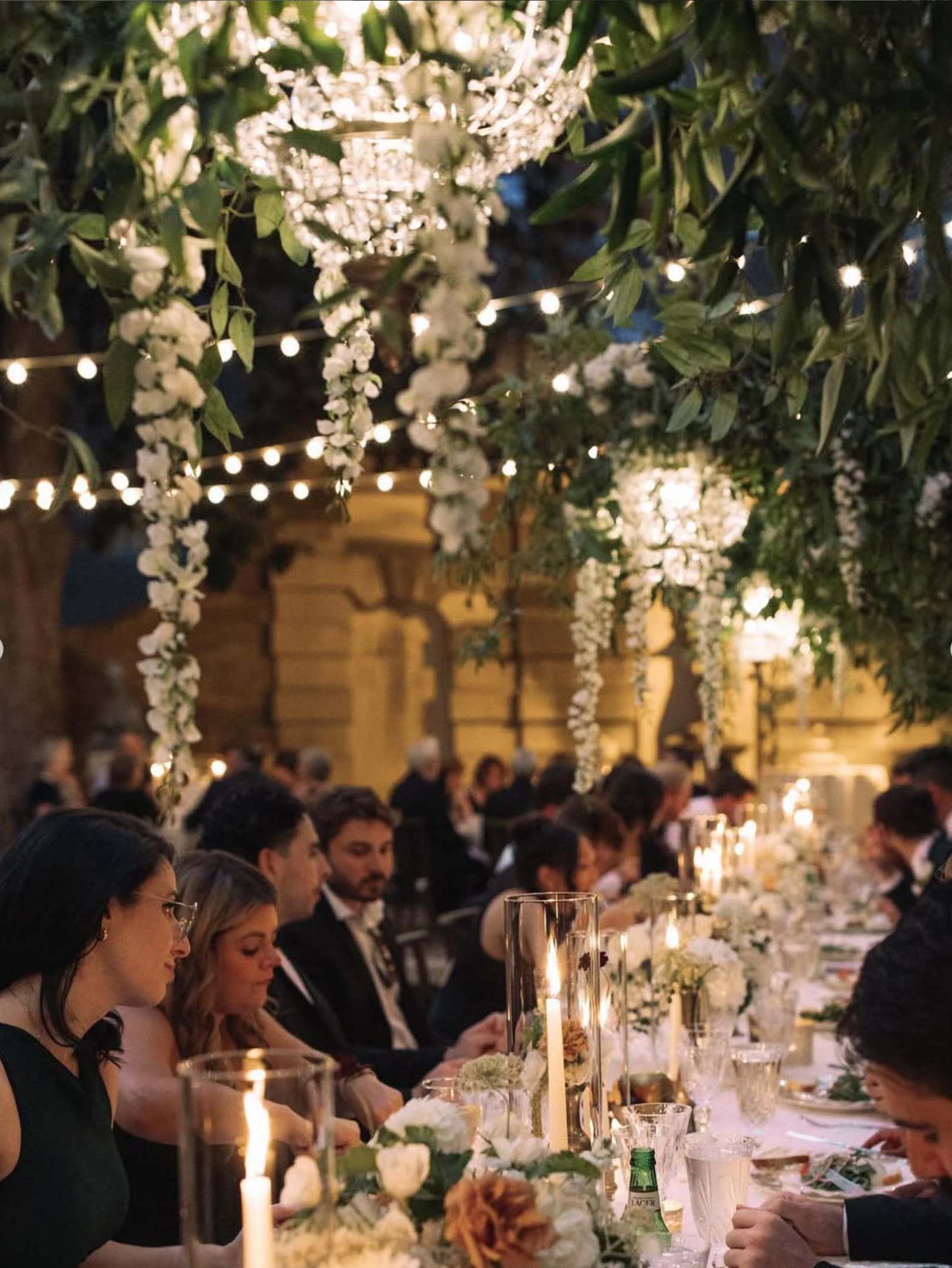 Outdoor dinner reception for a wedding—guests seated under string lights beside the Beaux Arts facade, with tall floral arches and candlelit tables