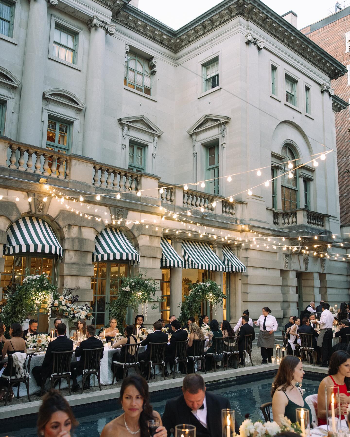 Elegant wedding reception featuring candlelit courtyard dining with string lights, floral arches, and striped awnings