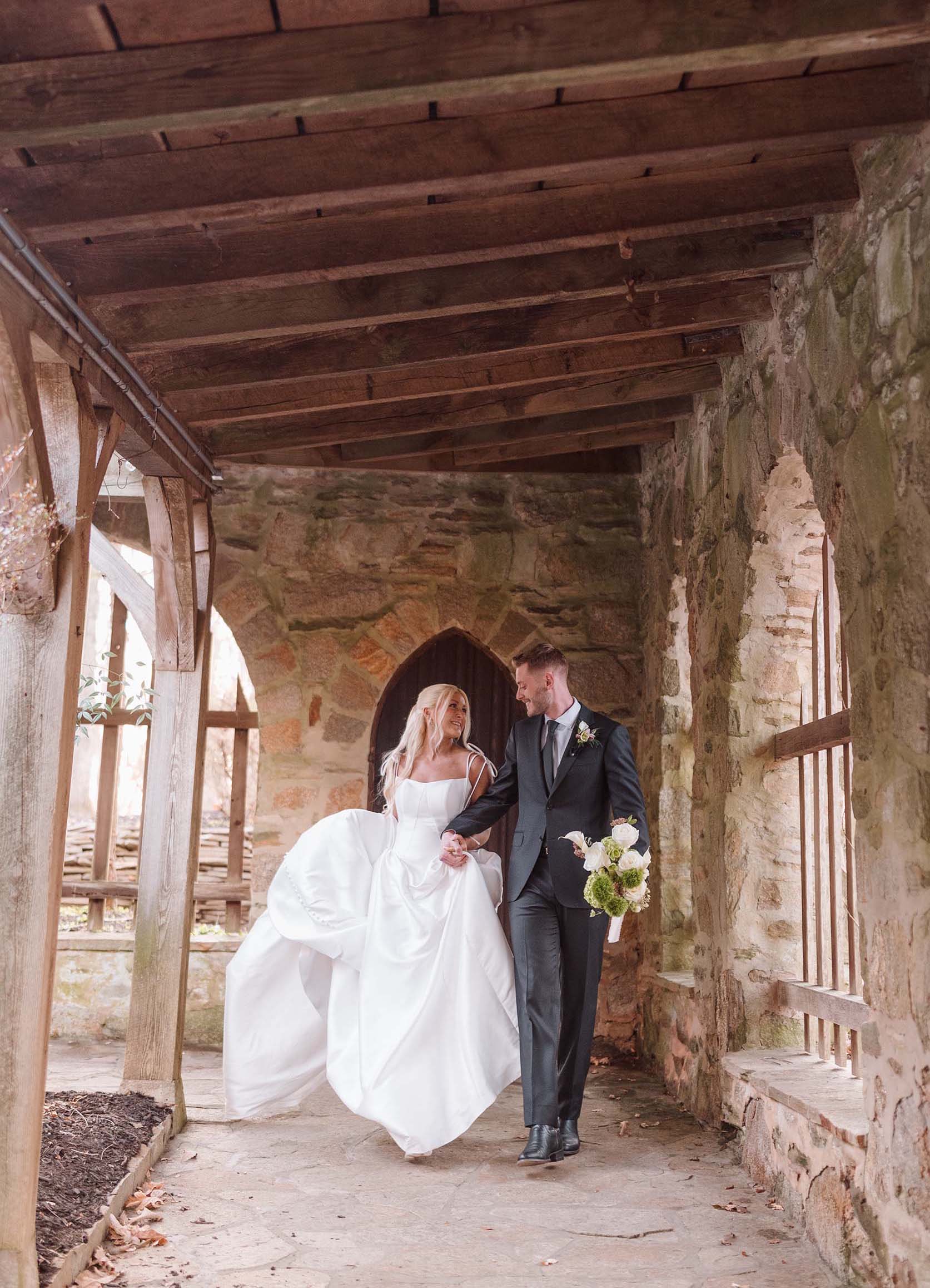 Newlyweds holding hands, walking through a castle’s stone corridor with joyful expressions.