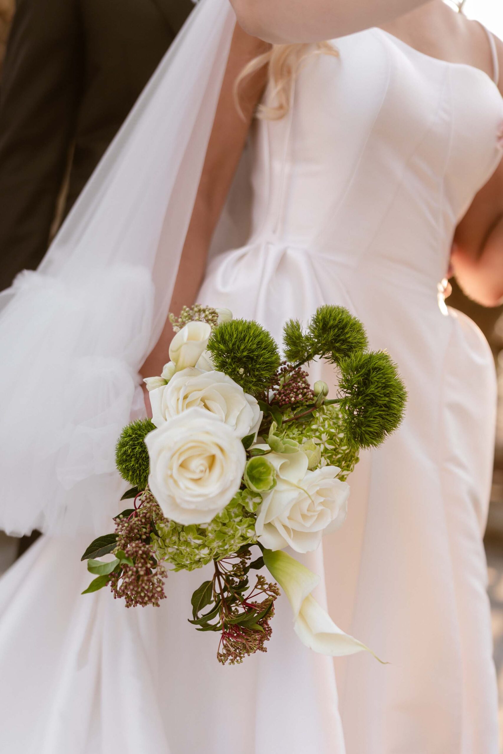Bride in satin gown holding a white and green wedding bouquet with roses, calla lilies, and textured florals.