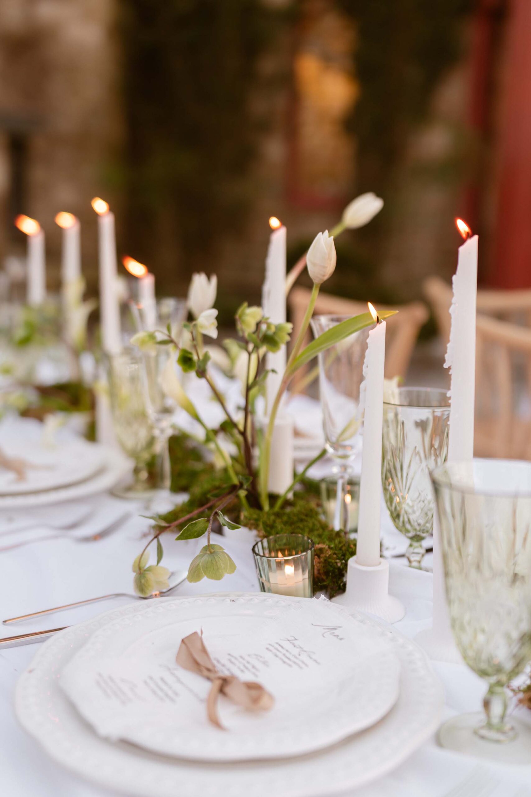 Elegant castle wedding tablescape with ivory calla lilies, glowing taper candles, crystal glassware, and a hand-torn menu tied with a golden ribbon atop white plates.