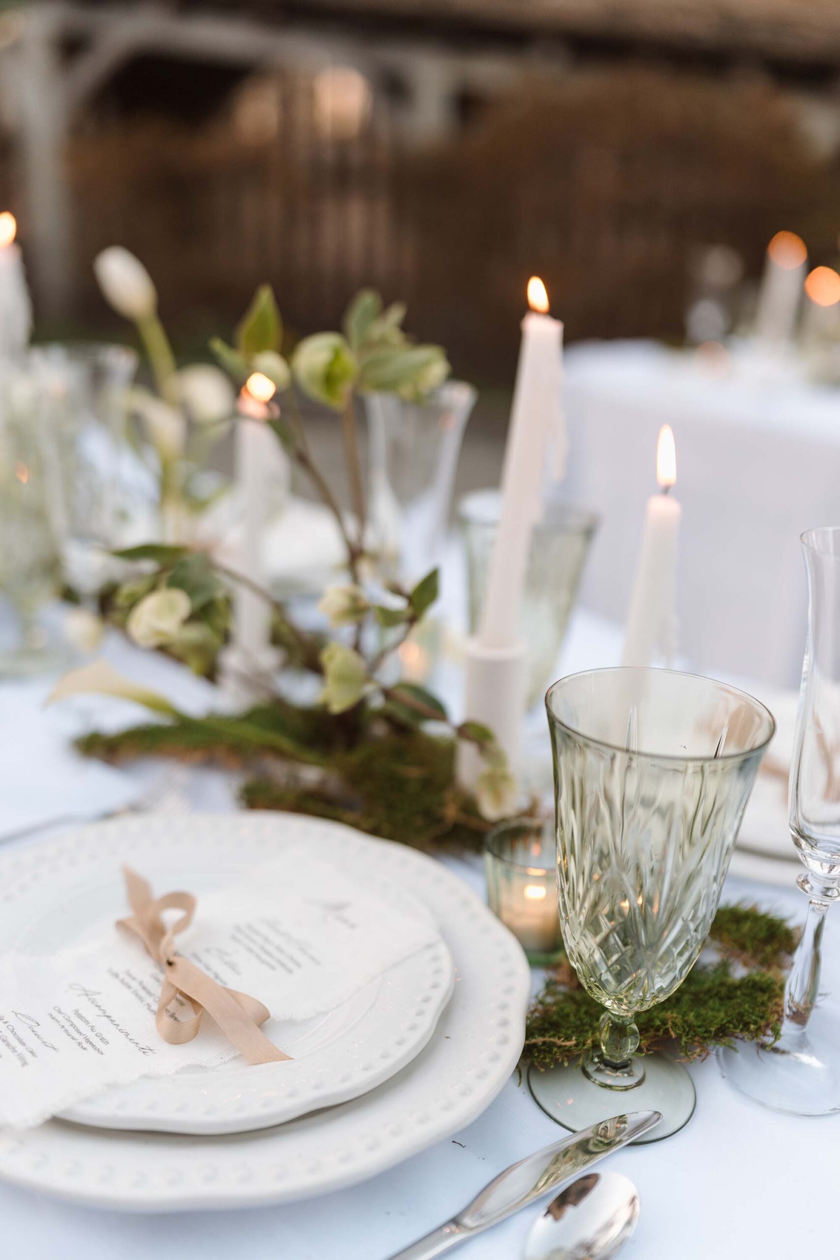 Romantic castle wedding place setting featuring white textured dinnerware, a taupe ribbon-tied menu, vintage green glassware, and flickering taper candles surrounded by greenery.