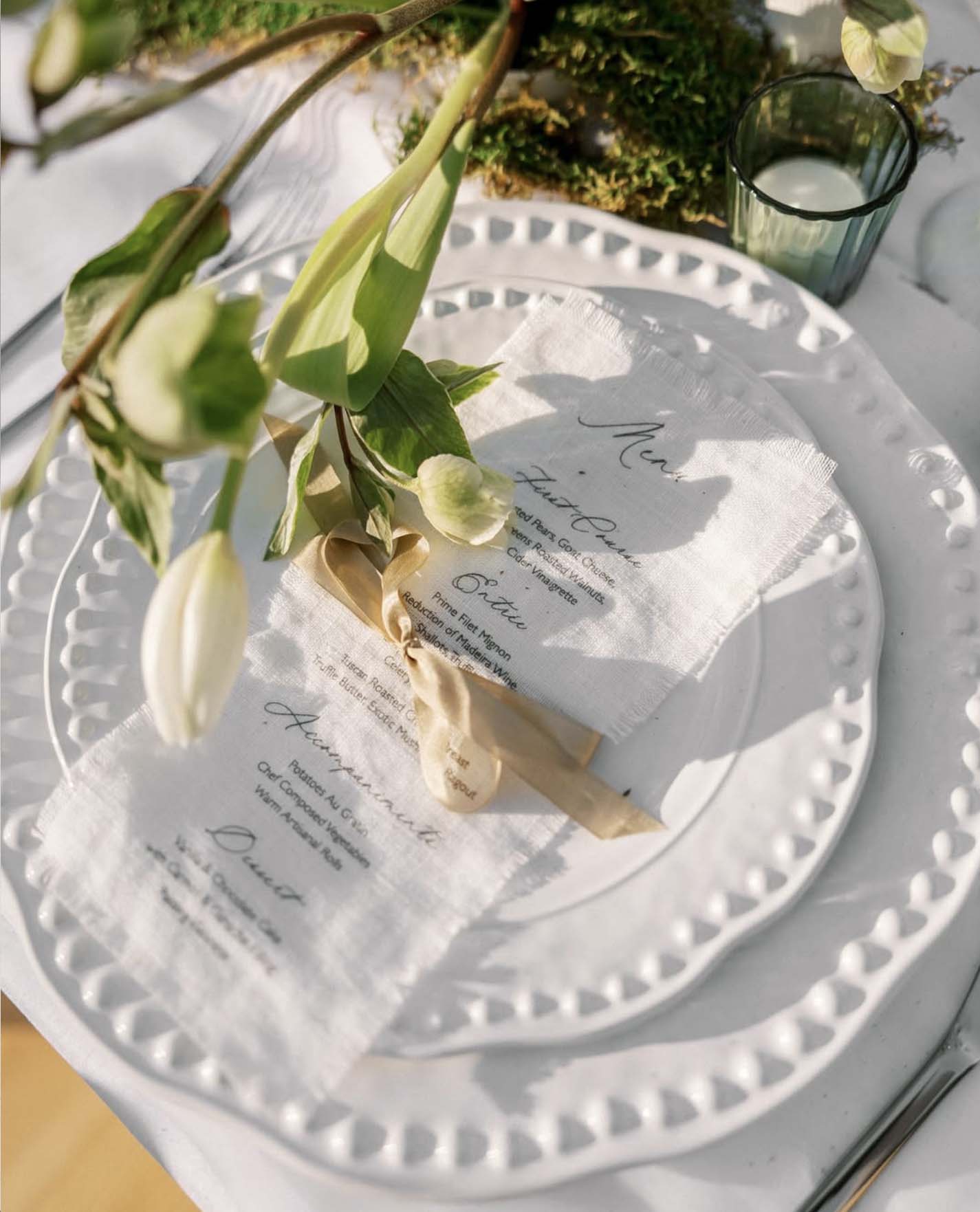 Closeup of linen menu tied with champagne ribbon on white plate with tulip centerpiece at The Cloisters Castle wedding reception