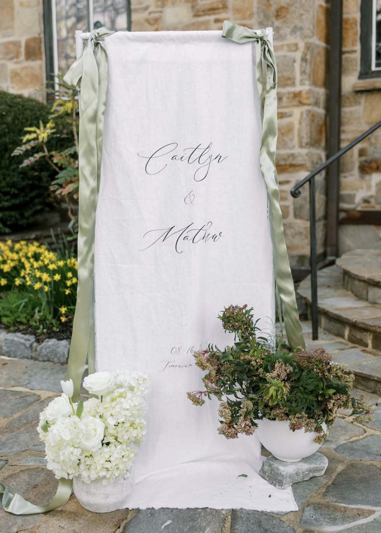 Custom linen wedding welcome sign with sage ribbon and white florals at The Cloisters Castle in Lutherville, Maryland