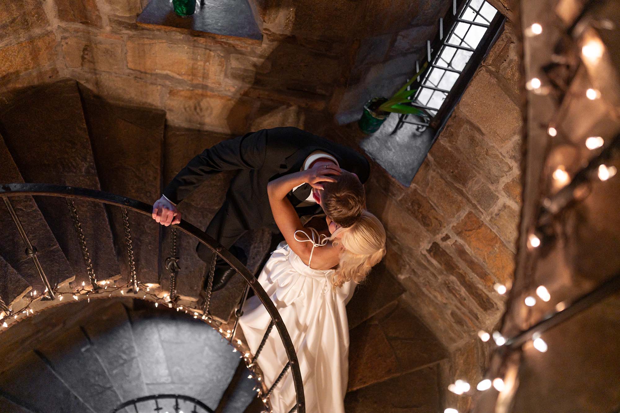 Bride and groom kissing on a spiral stone staircase lit by twinkling string lights at a castle wedding