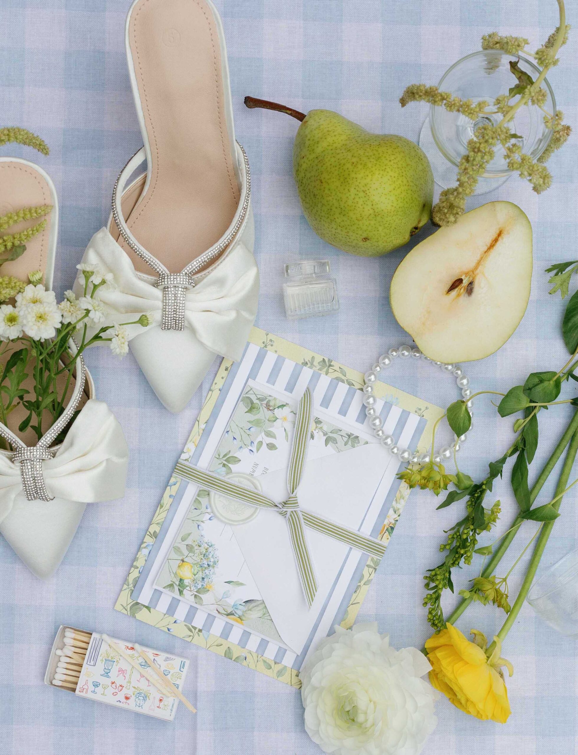 Elegant flatlay of a summer wedding invitation suite with ribbon wrap, pearl bracelet, satin heels, pears, yellow florals, and soft blue gingham linen.