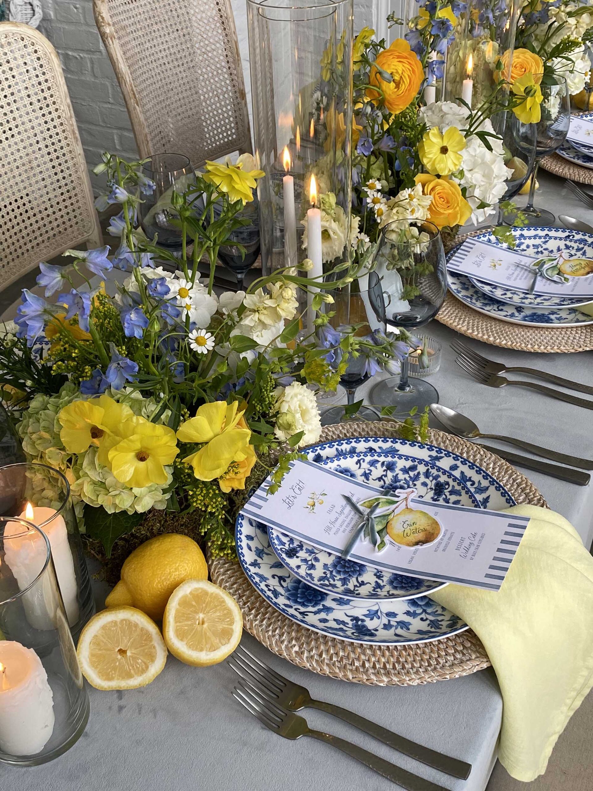 Vibrant yellow and blue wedding tablescape featuring lemon-themed menus, glass candle holders, wildflower centerpieces, and blue patterned plates.