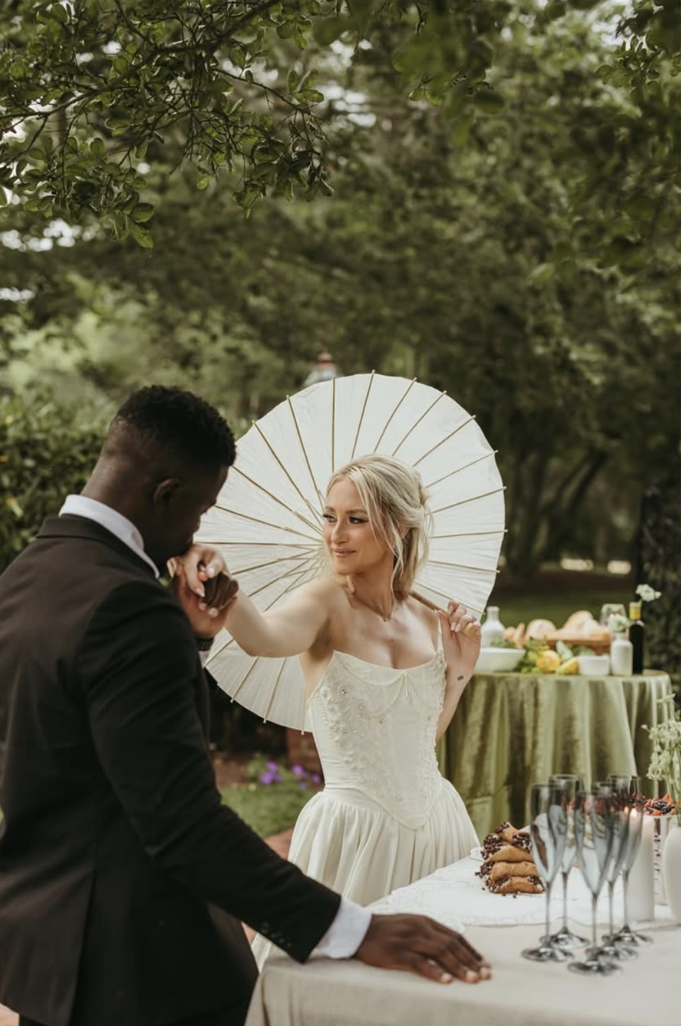 Groom kisses bride’s hand as she holds a parasol beside a dessert table set with cannolis and fresh fruit at Brittland Estates.