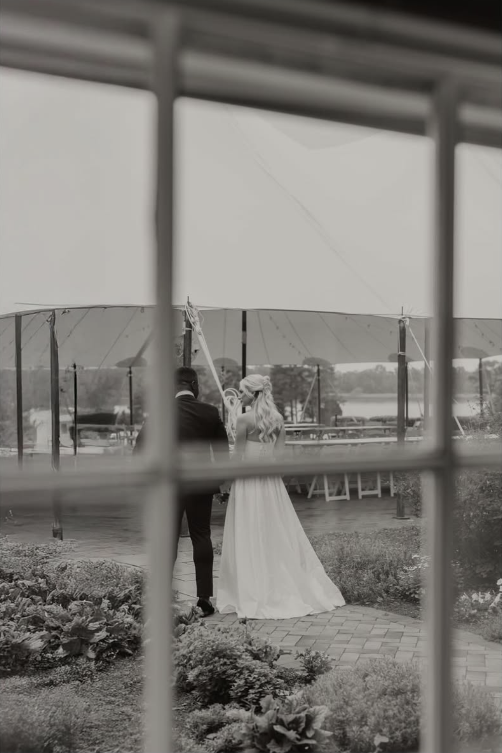 Black and white photo of newlyweds walking through the garden at Brittland Estates viewed through a window pane.
