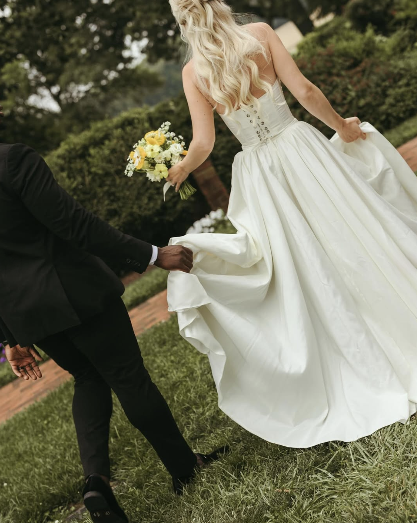 Groom holds bride’s train as she walks through garden path at Brittland Estates holding a yellow and white bouquet.