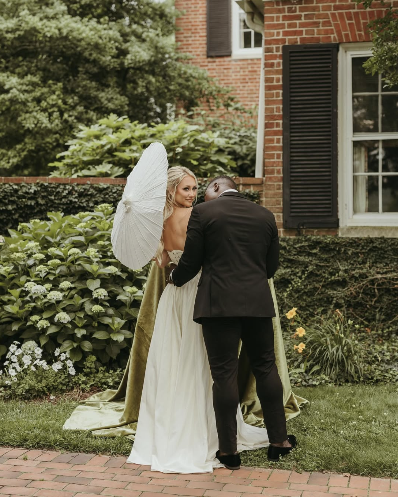 Bride with parasol glances back at the camera while walking arm in arm with groom beside ivy-covered brick wall at Brittland Estates.