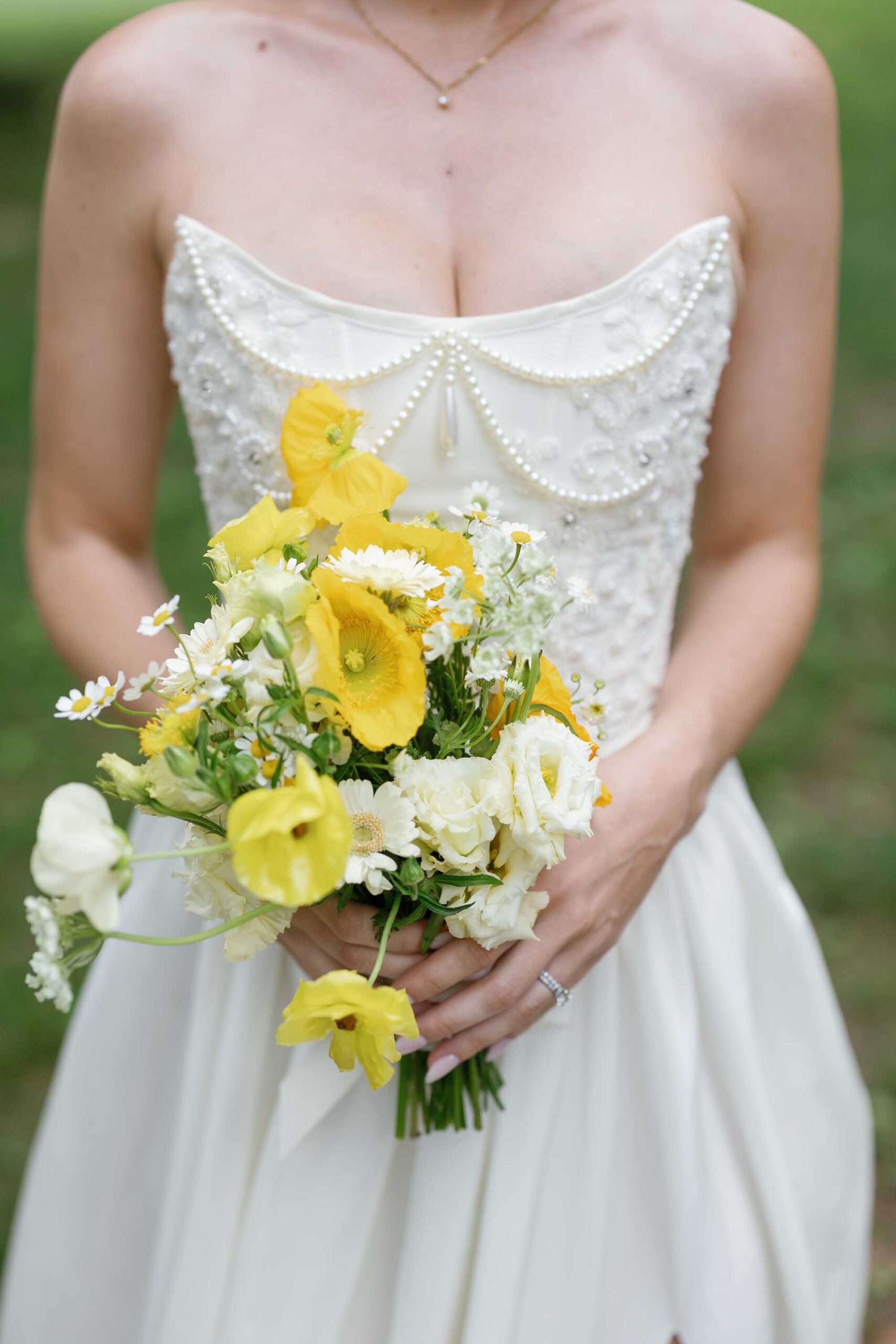 Close-up of a bride holding a yellow and white wildflower bouquet, featuring cheerful poppies and daisies, paired with an intricately beaded corset wedding gown.