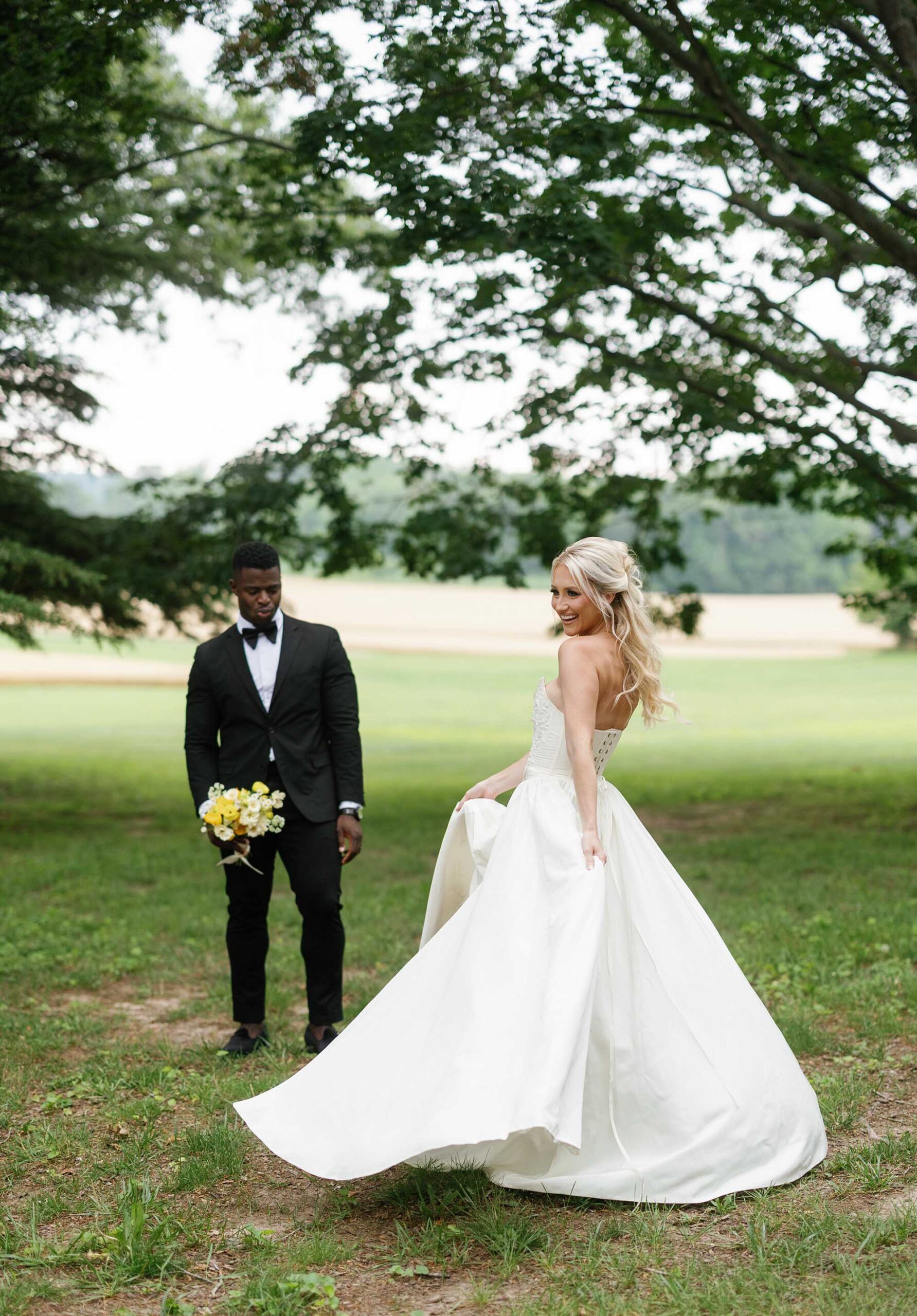 Bride twirling in her elegant strapless wedding gown under the trees at Brittland Estates, with groom in the background holding yellow and white bouquet.