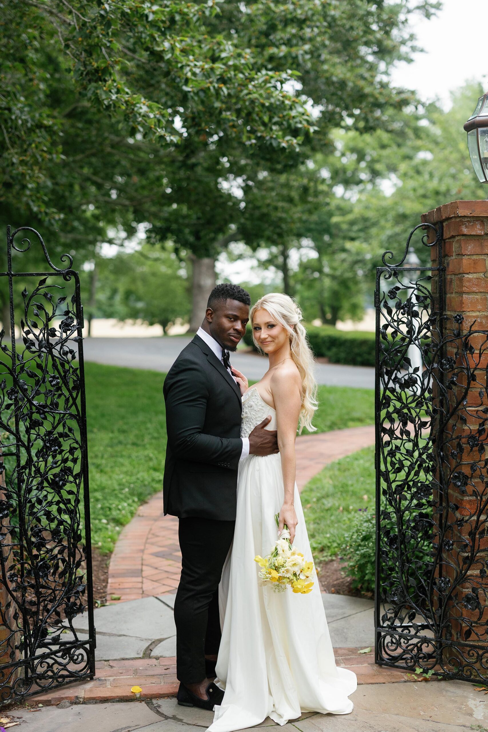 Bride and groom pose at ornate garden gates at Brittland Estates, holding yellow bouquet during Italian-inspired wedding shoot.