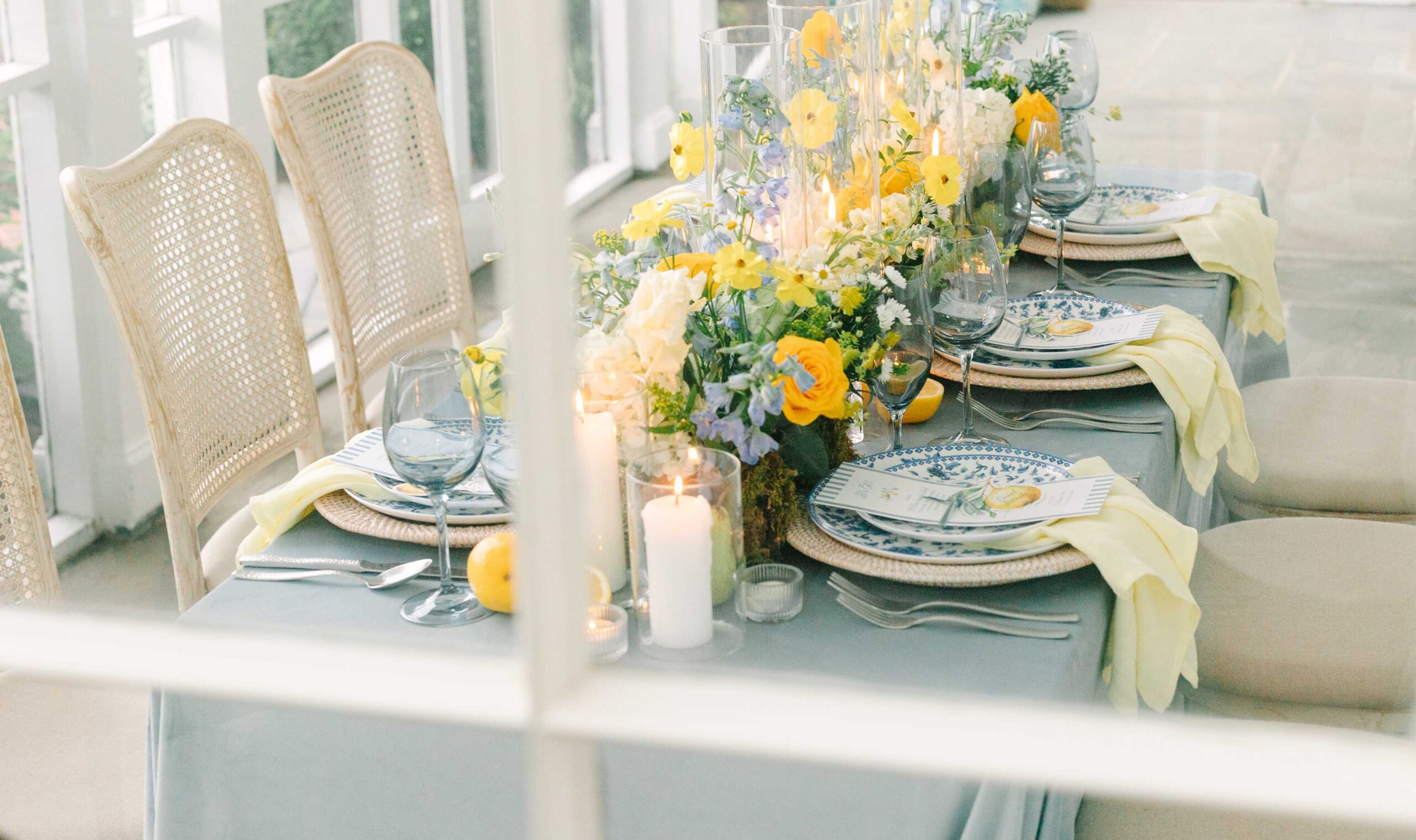 legant Mediterranean-inspired wedding tablescape with lemon yellow and cornflower blue florals, taper candles, and patterned china – captured through vintage-paned windows at Brittland Estates.