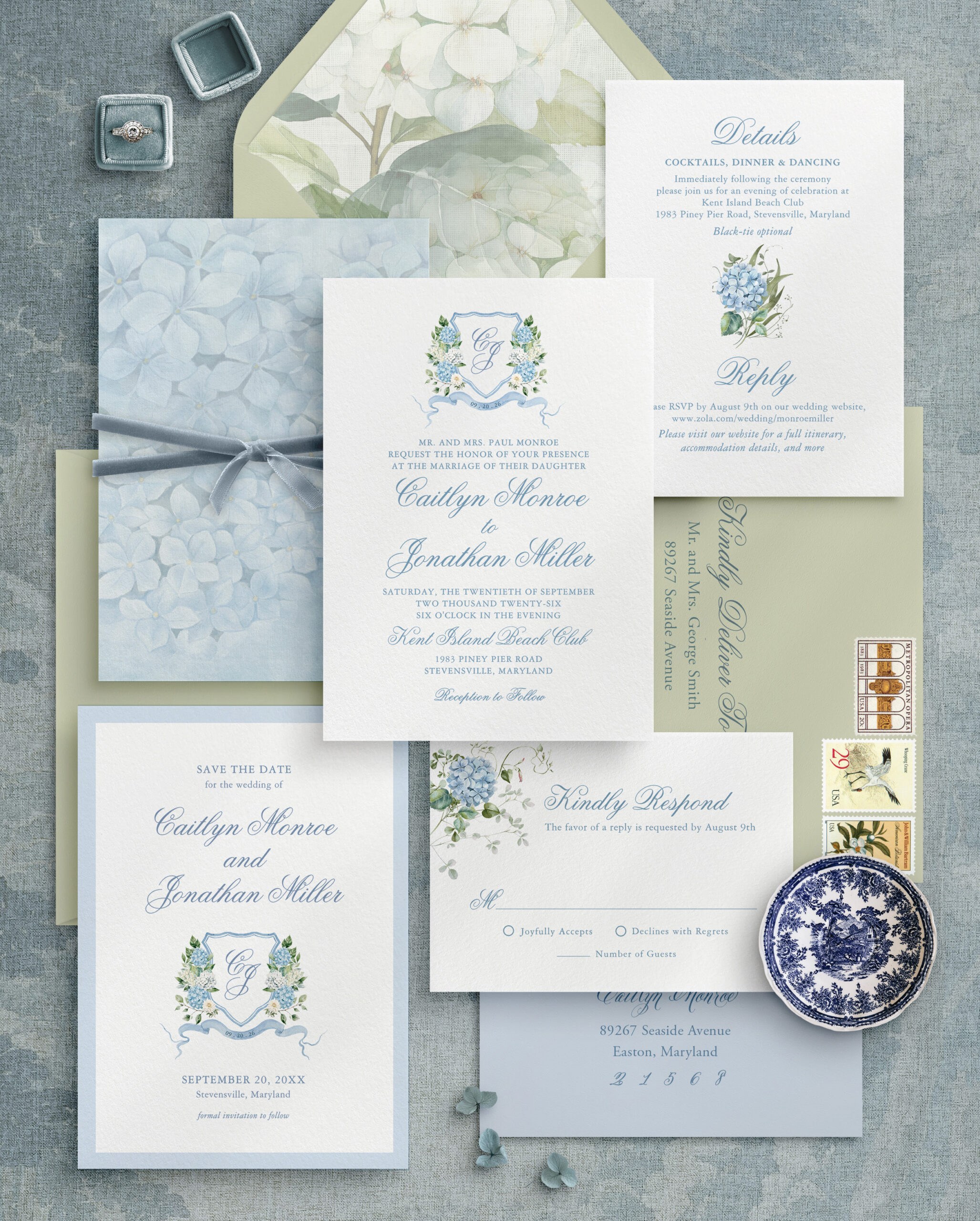Flat lay of blue hydrangea wedding invitations in a floral crest suite with save the date, RSVP card, envelope liner and details card on a blue linen background.