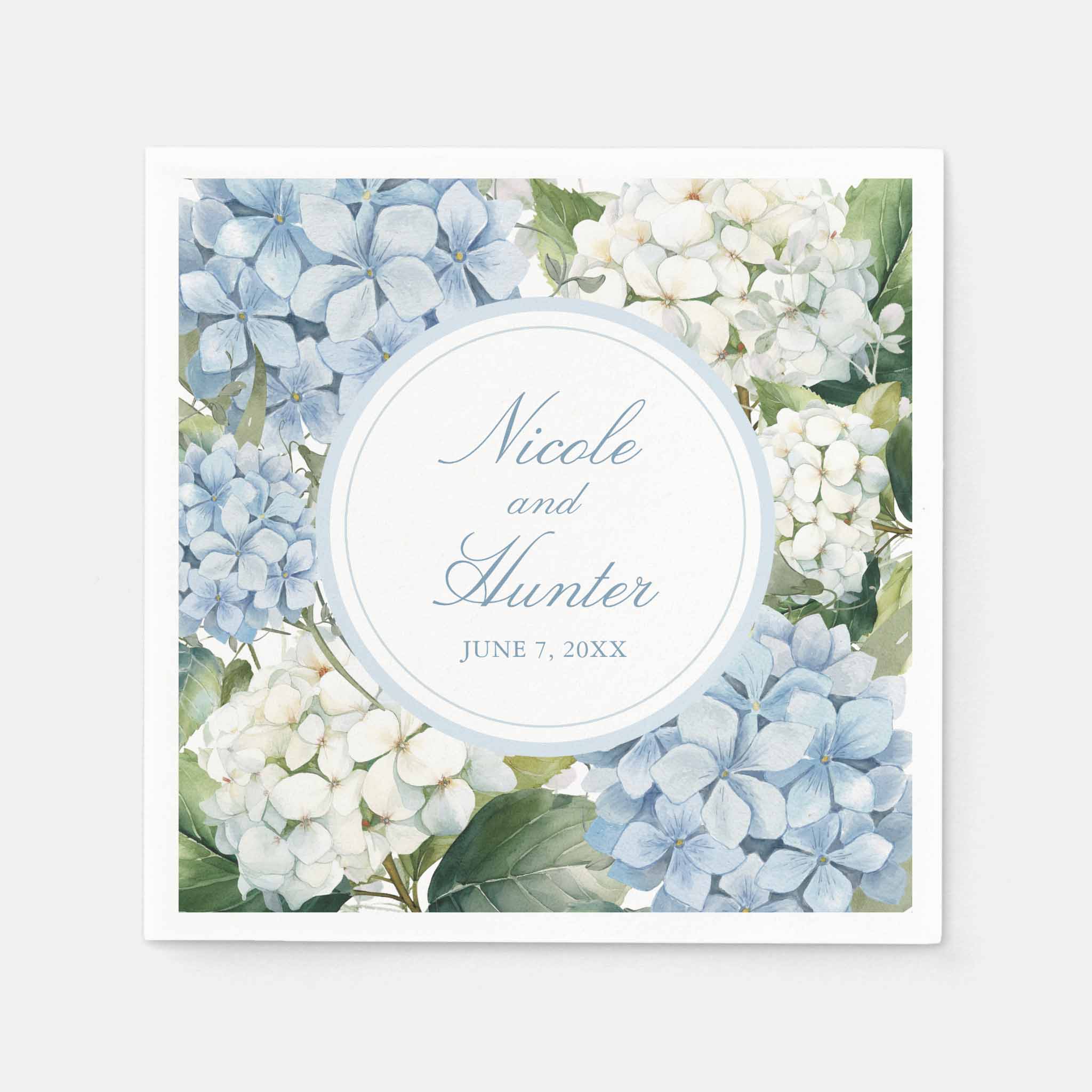 Personalized wedding cocktail napkin covered in blue and white hydrangea florals with circular frame showing couple’s names and wedding date.