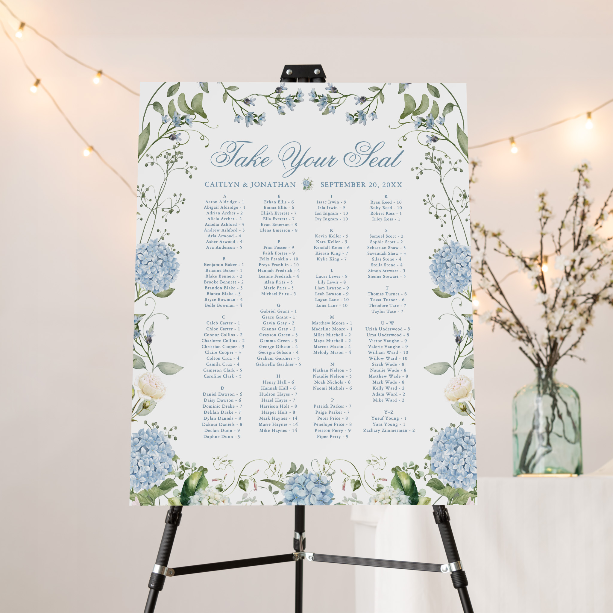 Take Your Seat wedding seating chart sign with blue hydrangea and greenery border listing guest names and table numbers alphabetically on easel.