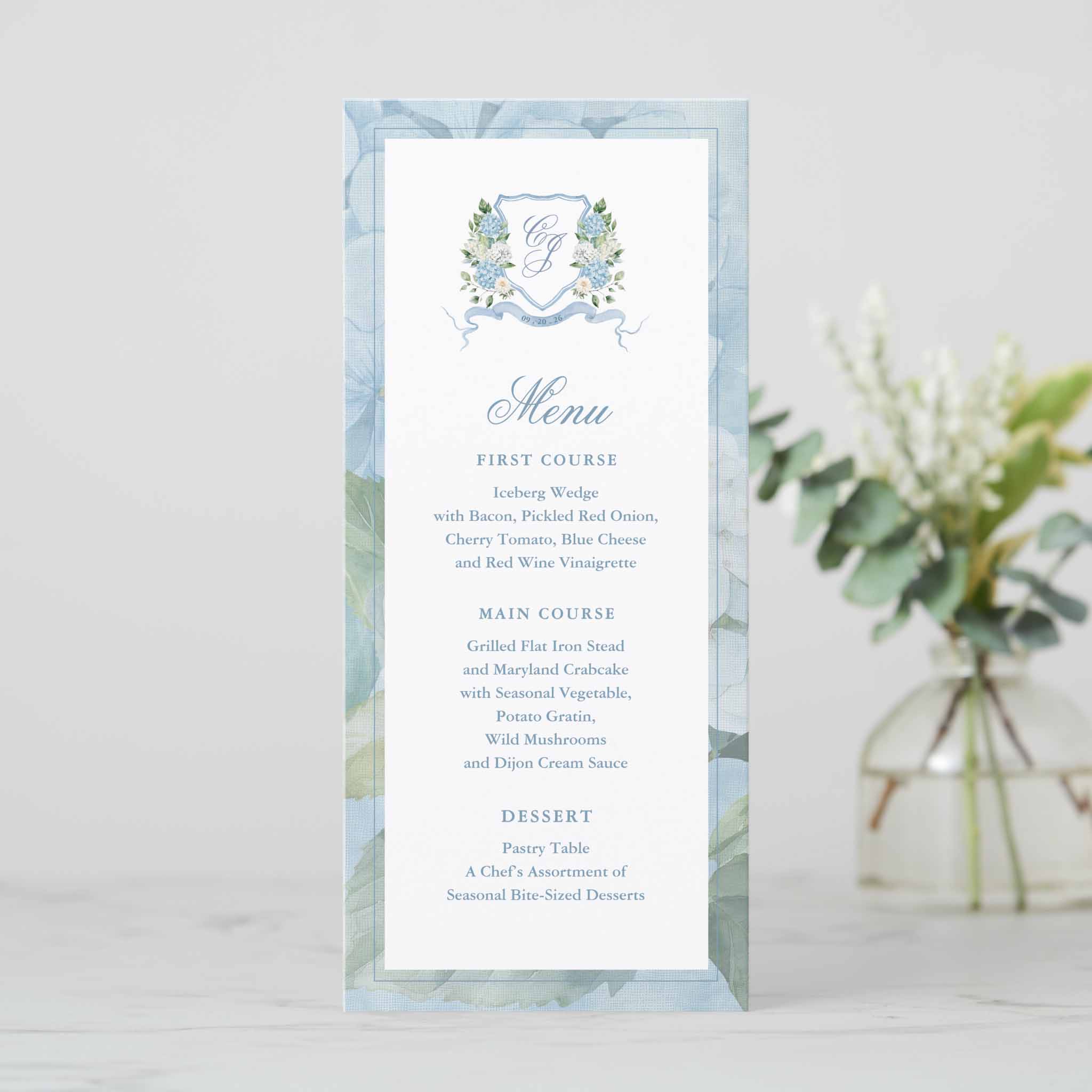 Tall wedding menu card framed with soft blue hydrangea and greenery border featuring floral crest monogram and classic menu wording.