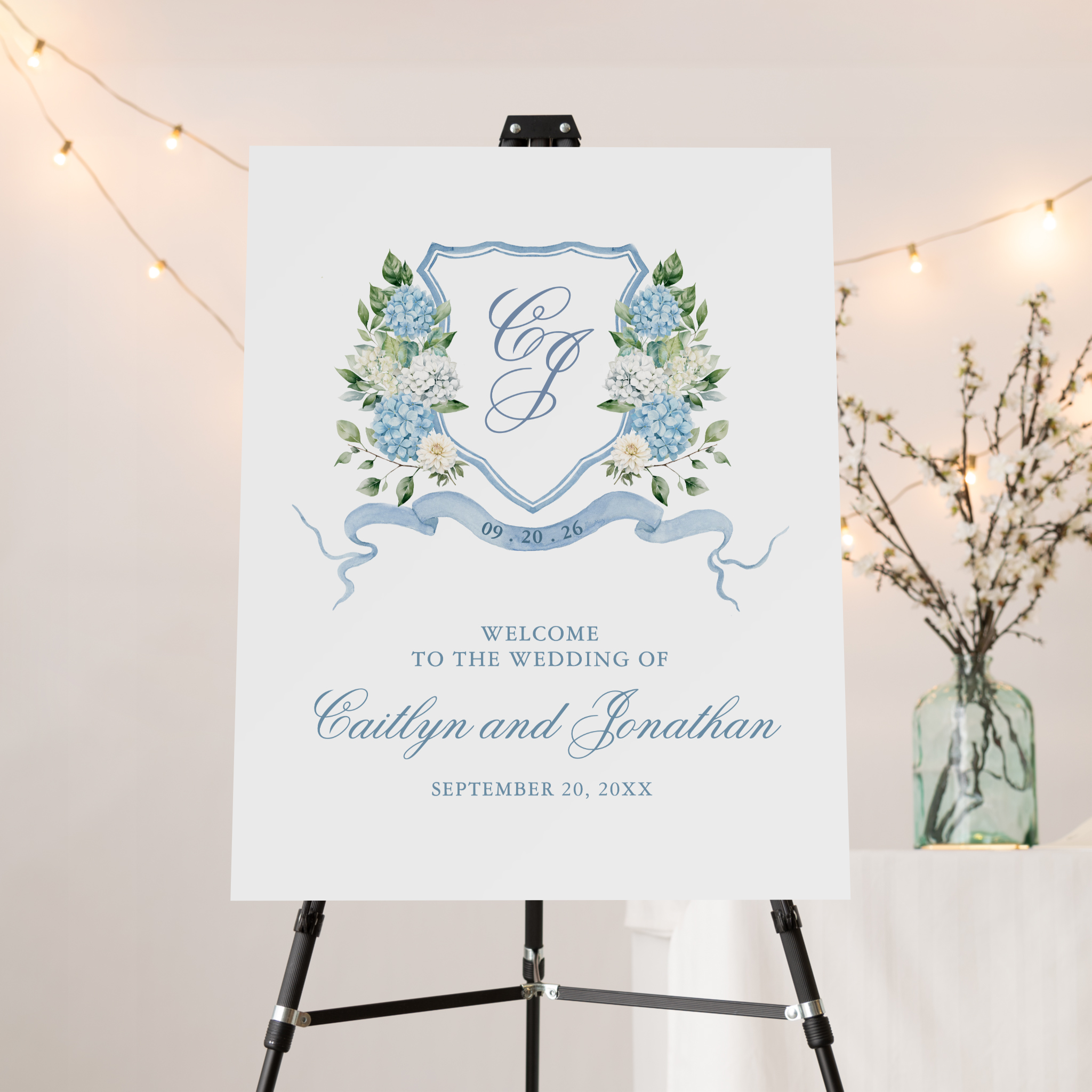 Wedding welcome sign on easel with blue hydrangea crest, script names and date, and soft ribbon detail for classic garden wedding.