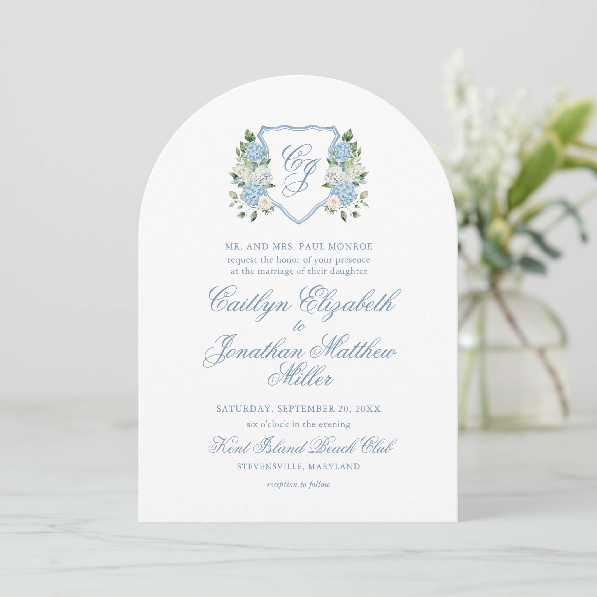 Arch shaped blue hydrangea floral crest wedding invitation featuring monogram shield and romantic script lettering.