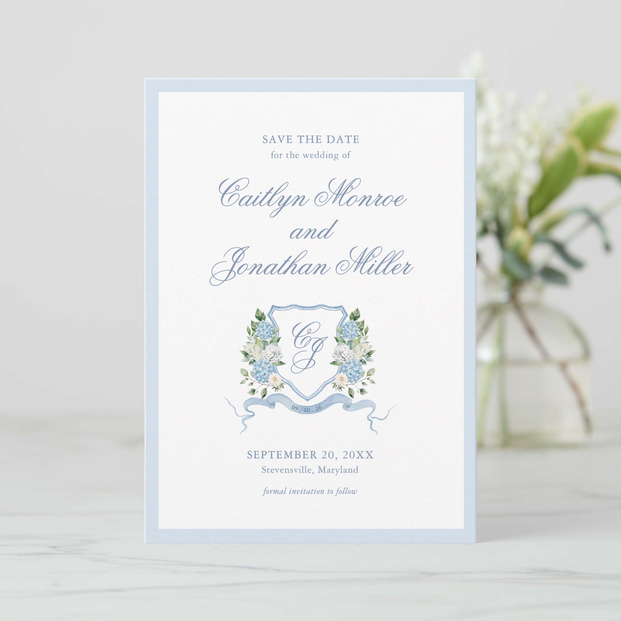 Blue hydrangea floral crest save the date card with romantic script text on white card stock styled on a light table.