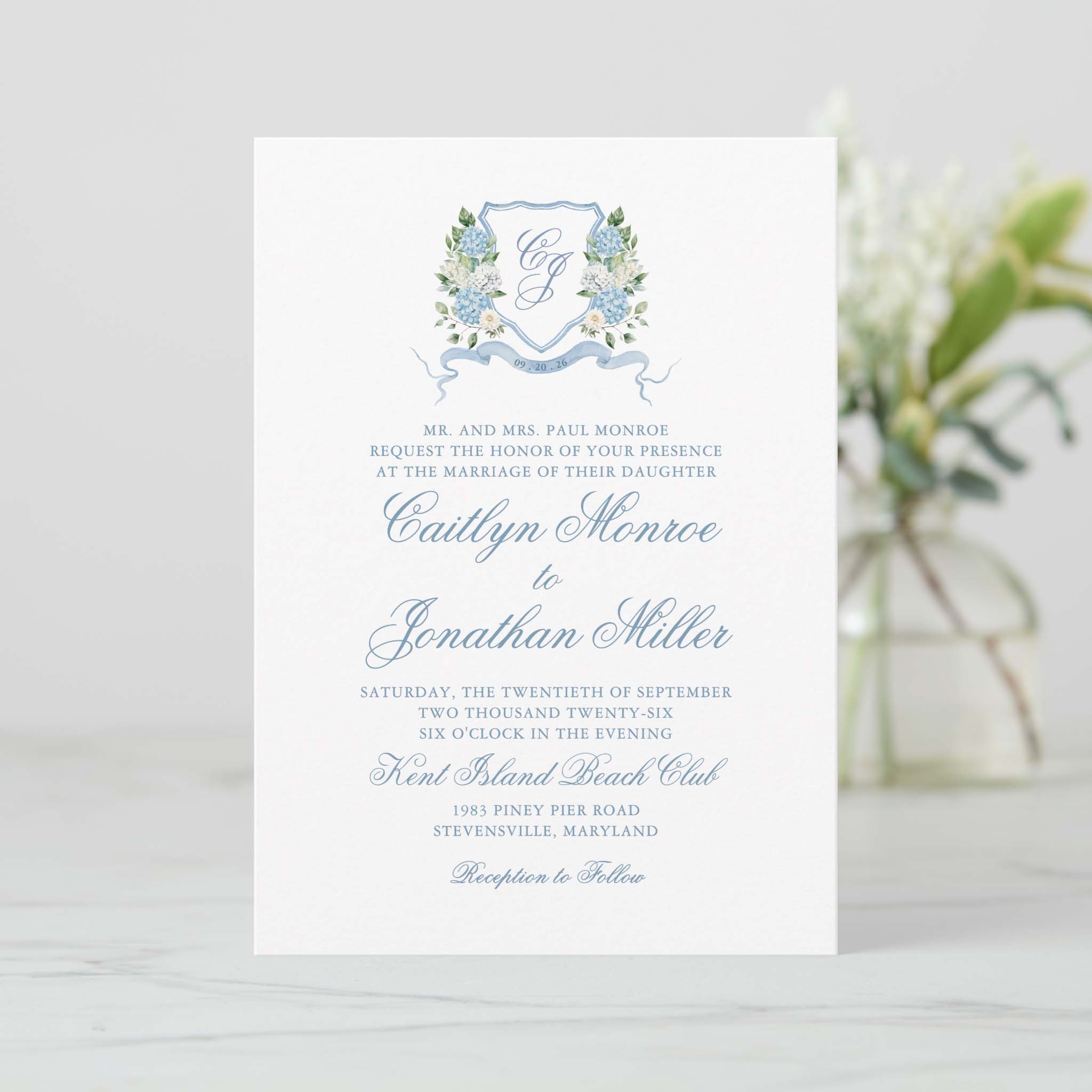 Blue hydrangea floral crest wedding invitation with monogram shield, ribbon banner and elegant script names.