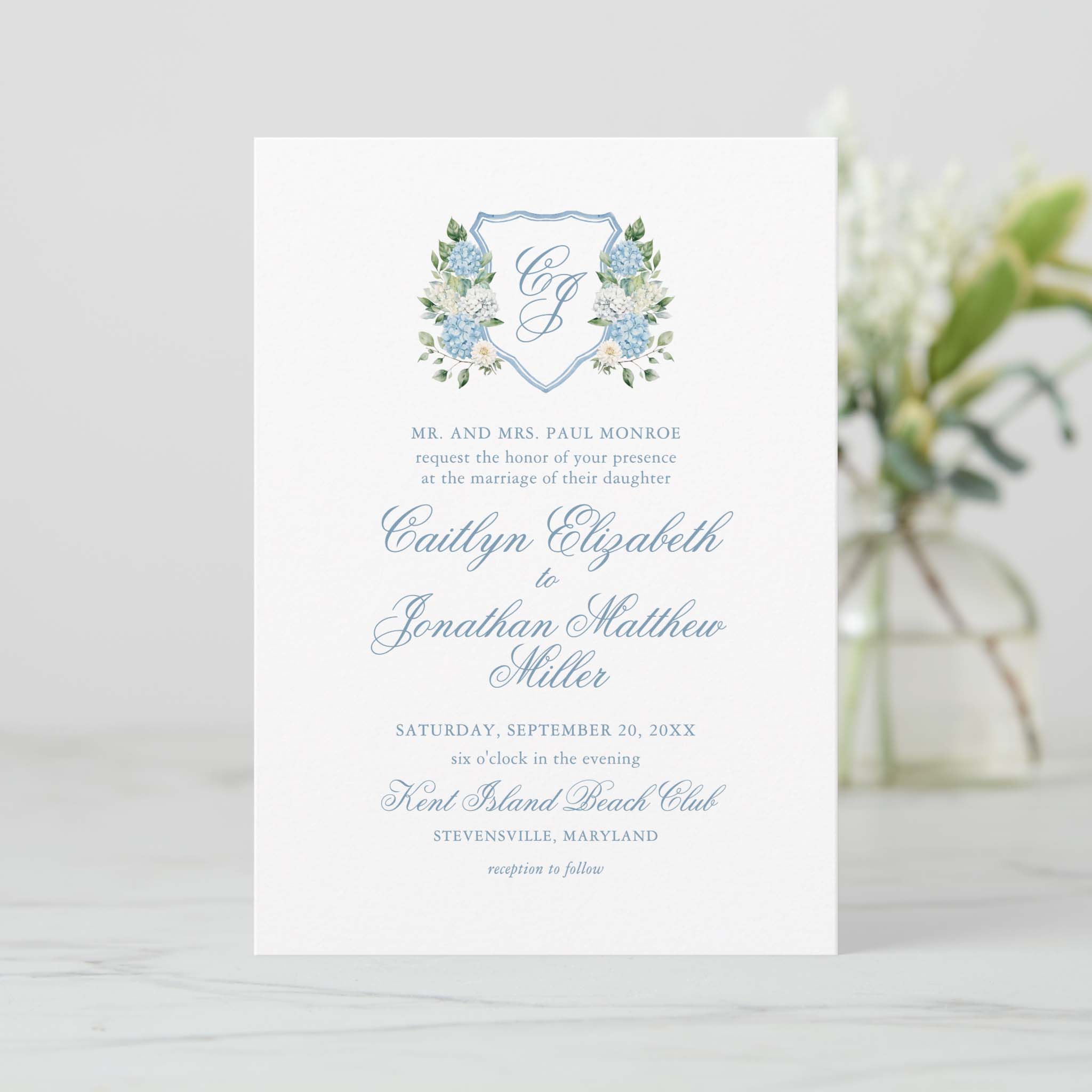 Classic blue hydrangea floral crest wedding invitation with monogram shield and script typography on white card stock.