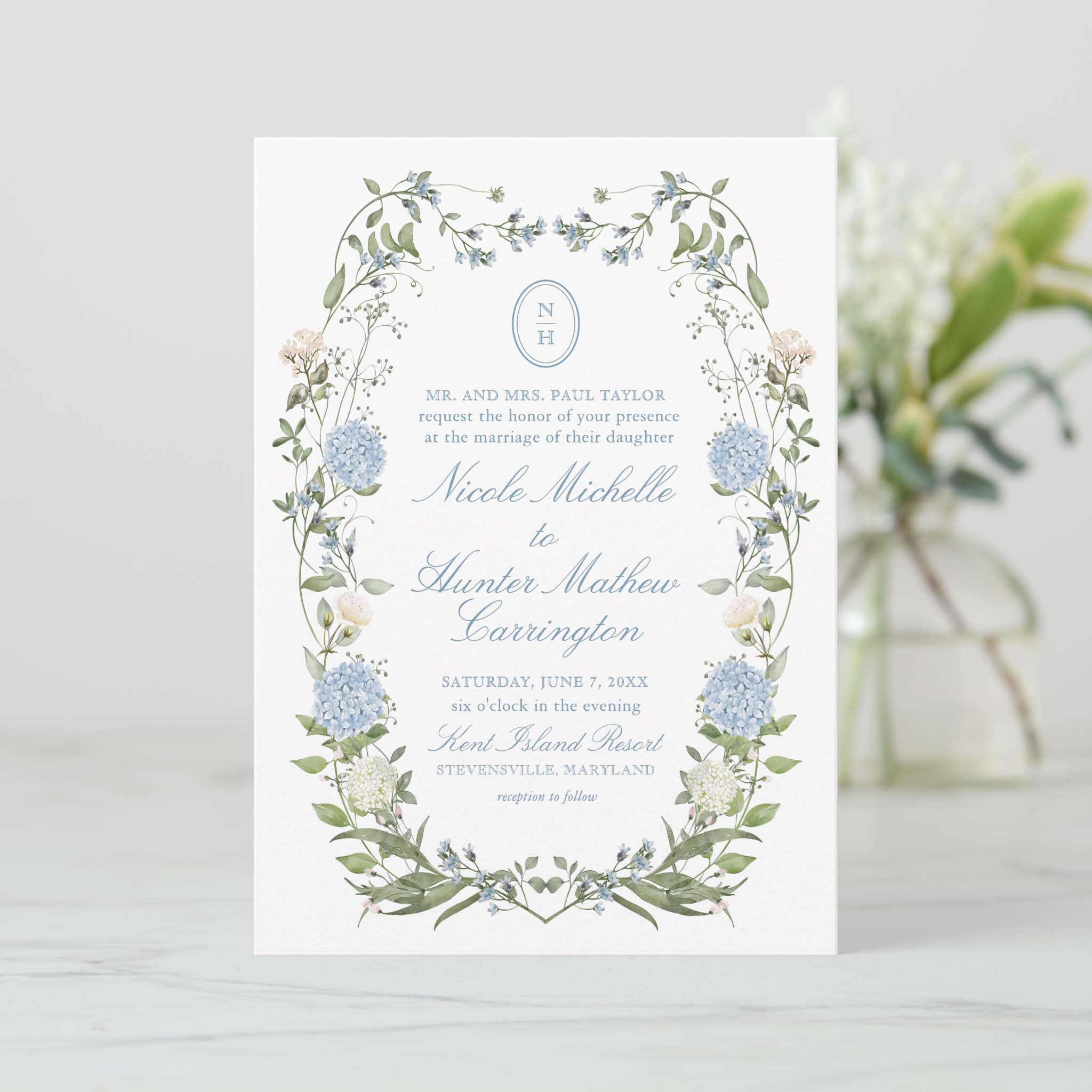 Dusty blue hydrangea wedding invitation with delicate greenery border and round monogram at the top on white background.