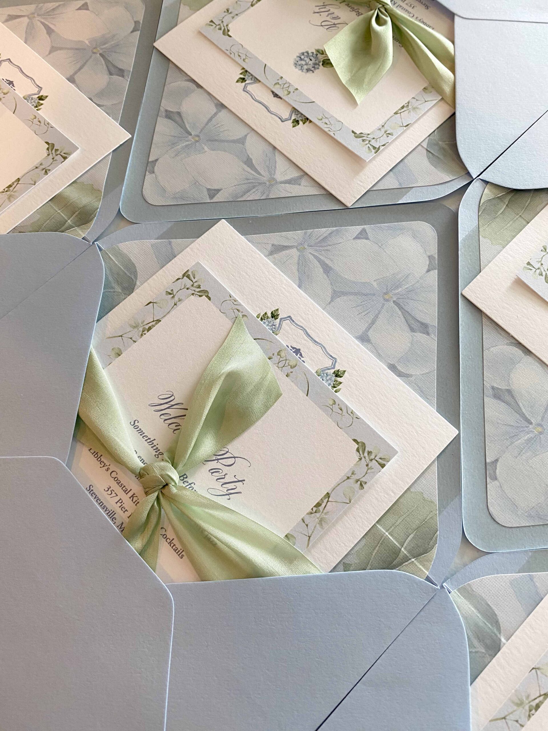 Stack of blue hydrangea wedding invitation suites tucked into light blue envelopes with floral liners and tied with soft green ribbon.
